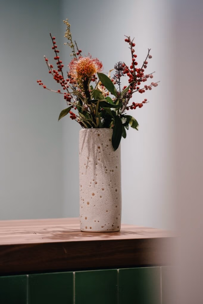 MORGEN & Friends Raumdetail mit Keramikvase, Blumen und ruhiger Dekoration auf Holzoberfläche.